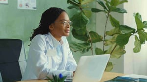 Young female medical professional with glasses sitting at a desk in front of a laptop looking out into the distance